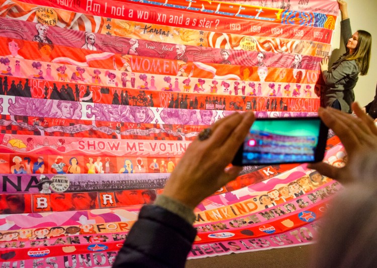 Amie Kennedy holds up part of the "Her Flag" project Saturday after Marilyn Artus added Kennedy's Maine stripe to the bottom during an event at the Maine State Museum in the Augusta.