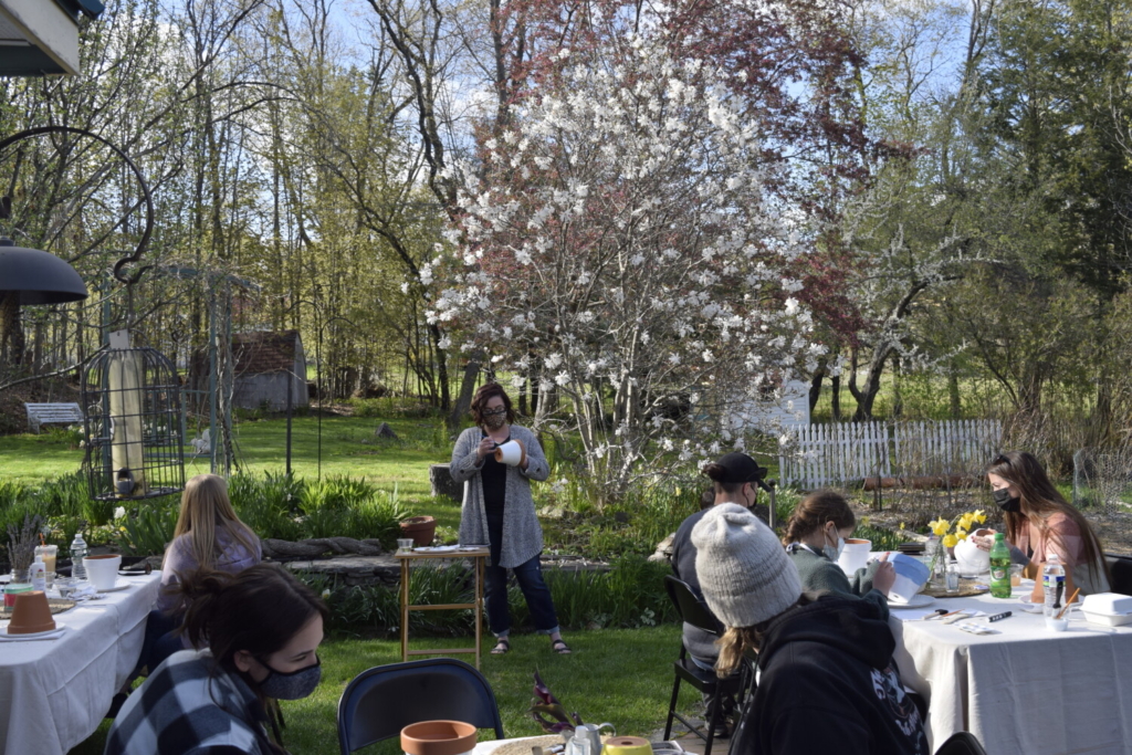 Five adults and two children sit, in a garden, at tables covered in art supplies and flower pots while a woman stands in the background painting a flower pot.