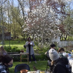 Five adults and two children sit, in a garden, at tables covered in art supplies and flower pots while a woman stands in the background painting a flower pot.